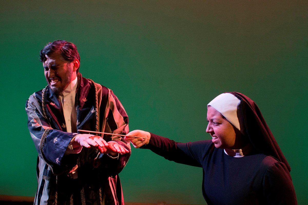 A turns his head and grimaces while a nun slaps his hands with a ruler.