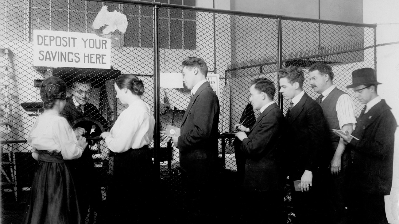 Two women and five men in old fashioned clothing stand before a desk.