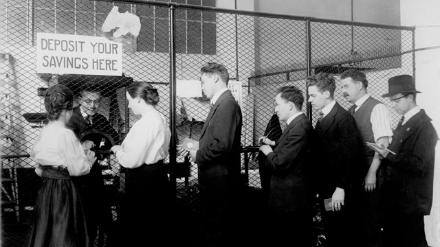 Two women and five men in old fashioned clothing stand before a desk.