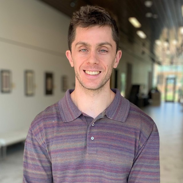 White man with short brown hair and stubble wearing a purple shirt.