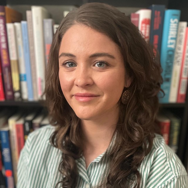 A white woman with long brown hair posing in front of a bookshelf