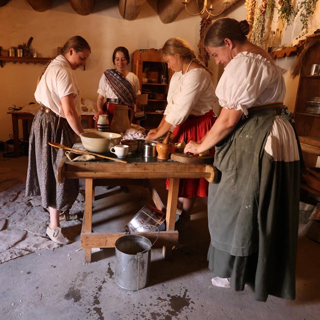 Four women dressed in 19th century clothing stand around a table in an adobe kitchen