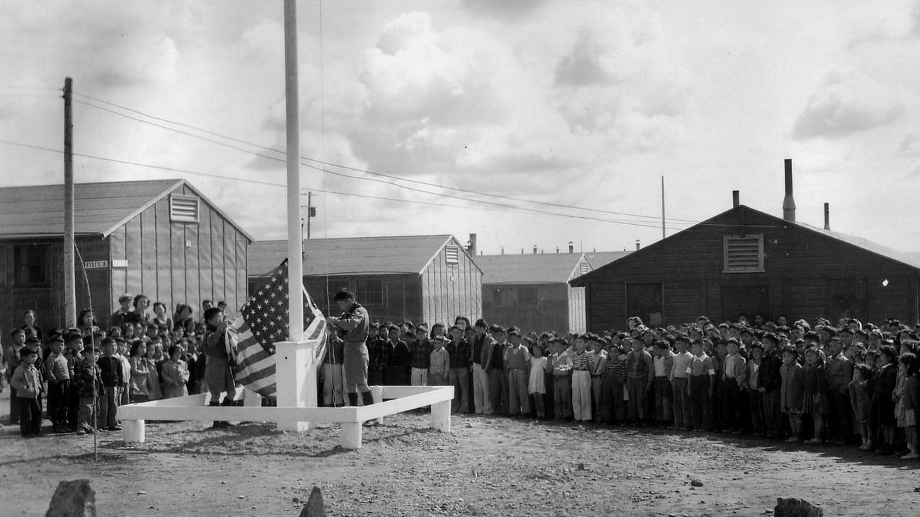 A large crowd of people watch two people holding the flag at a flagpole in front of barracks.