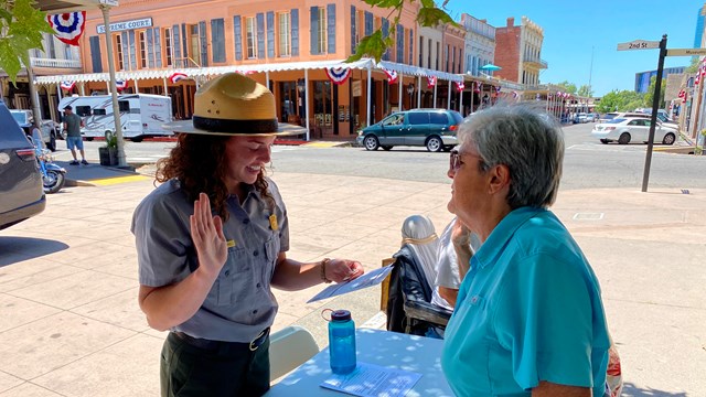 A park ranger holds up their hand and reads off a card to a person at a table outside.