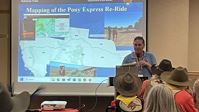 A person stands in front of a screen presentation, speaking to a group of peoplel