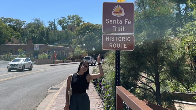A person stands underneath a brown sign on the side of the street.
