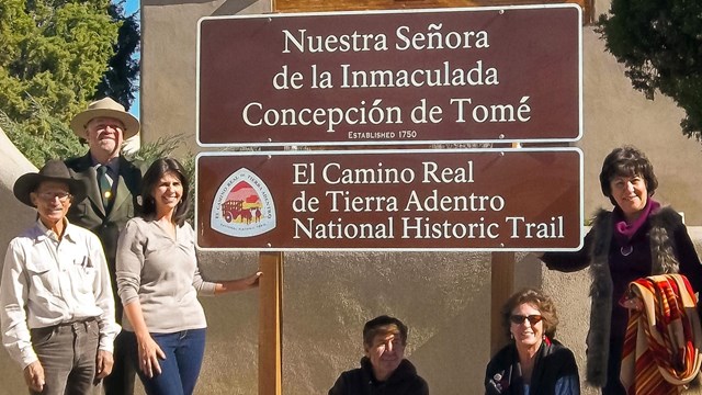 People standing and sitting next to a brown sign with white text in English and Spanish