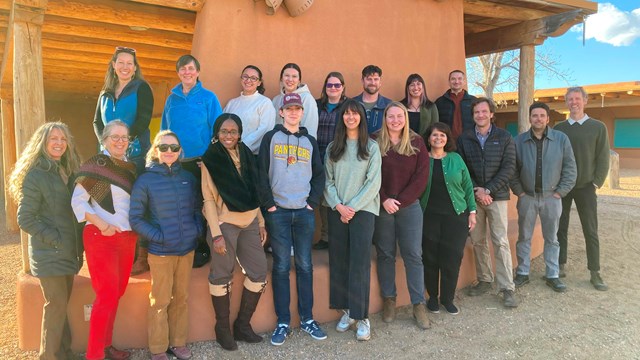 A group of about 20 people stand for a photo in a desert setting next to a wall.