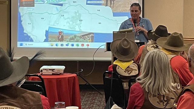 A person stands in a front screen, presenting to a group of people in chairs.