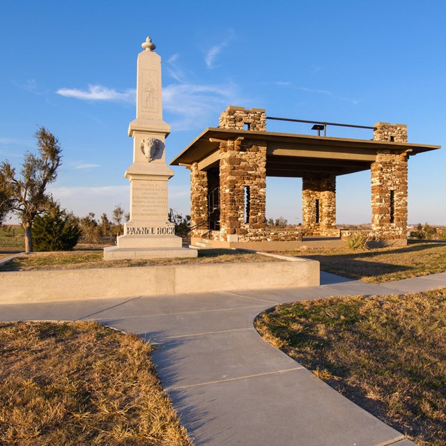 Tall, carved stone monument next to a covered shelter with columns or rock on top of a hill.
