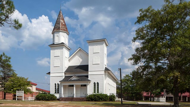 A neat white church with two rectangular turrets, one with a shingled roof, is surrounded by lawn.