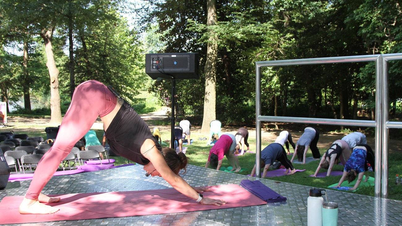 A yoga instructor leads a yoga class in a downward dog pose from an elevated stage