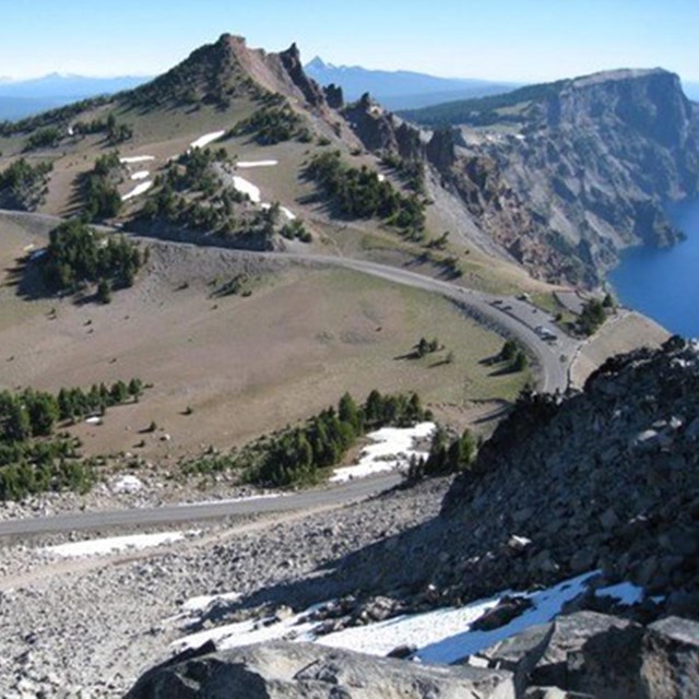 a road winds along a hill crest beside the cliffs of a deep blue lake