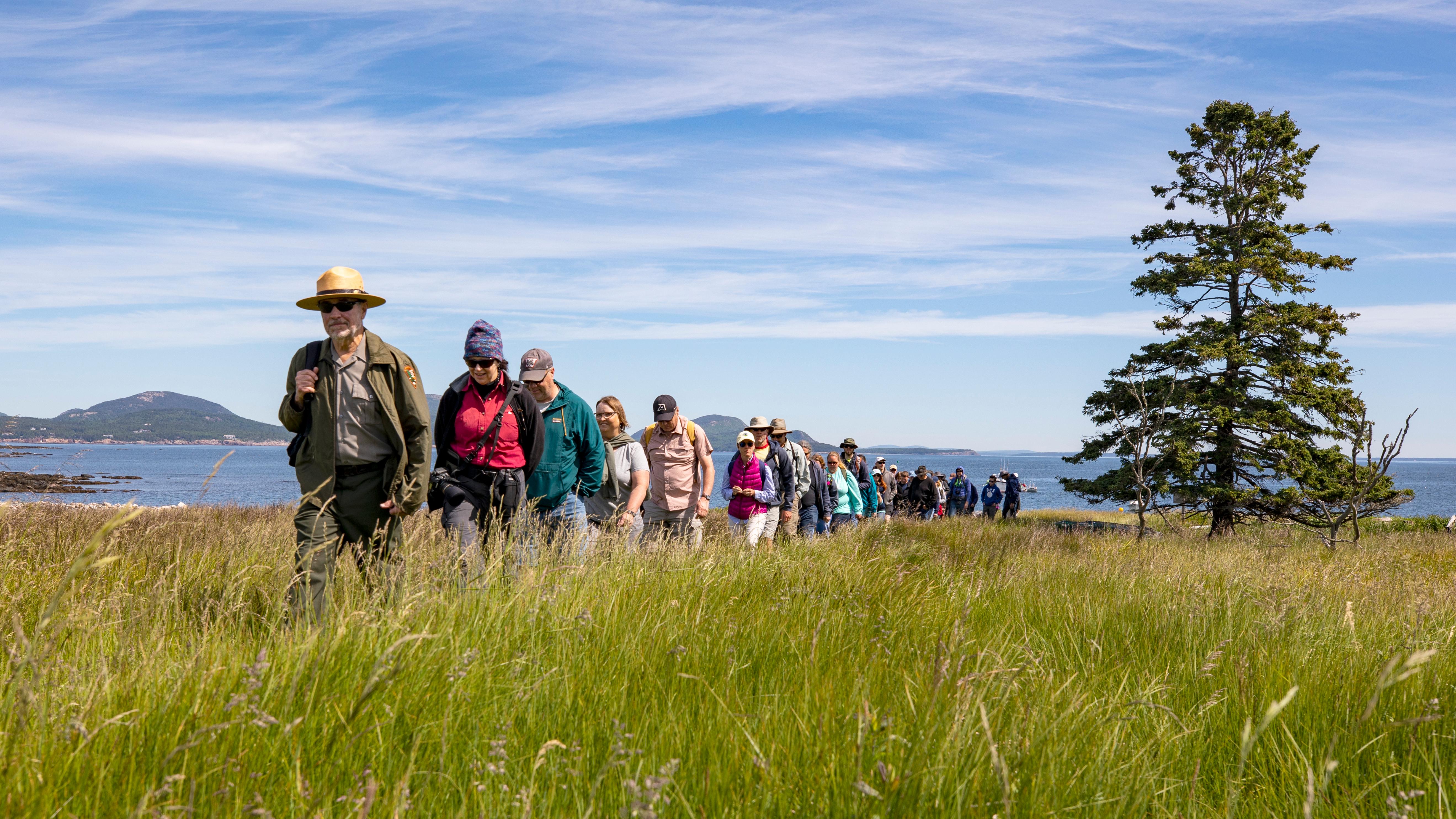 Ranger leads visitors walking single file through a field with a coastline and mountain in distance