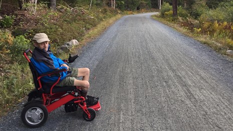 A person on an electric wheelchair gestures down a wide gravel path framed by trees