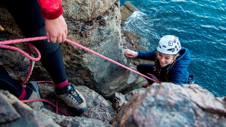 A climber in a white helmet climbs rocky cliff over water, belayed by someone mostly out of view