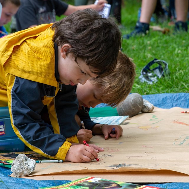 Two children draw with crayons on a large sheet of paper on tarp on grass