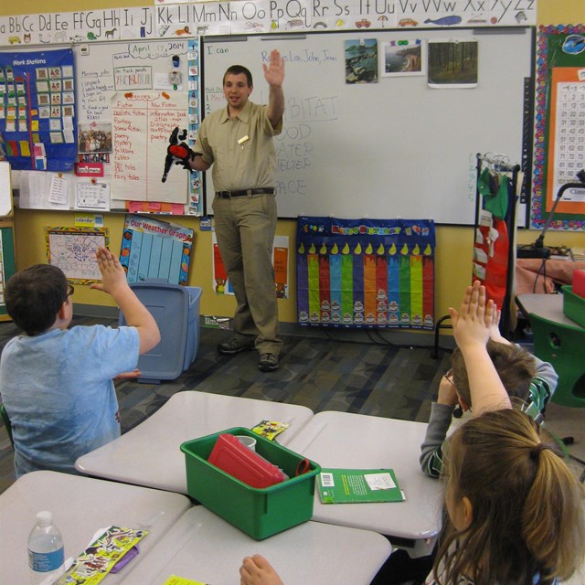 Man in uniform holding puppet in a classroom while students raise hands. 