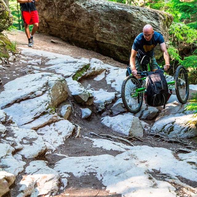 man on a prone hand-cycle, hiking on a rocky treed trail.  