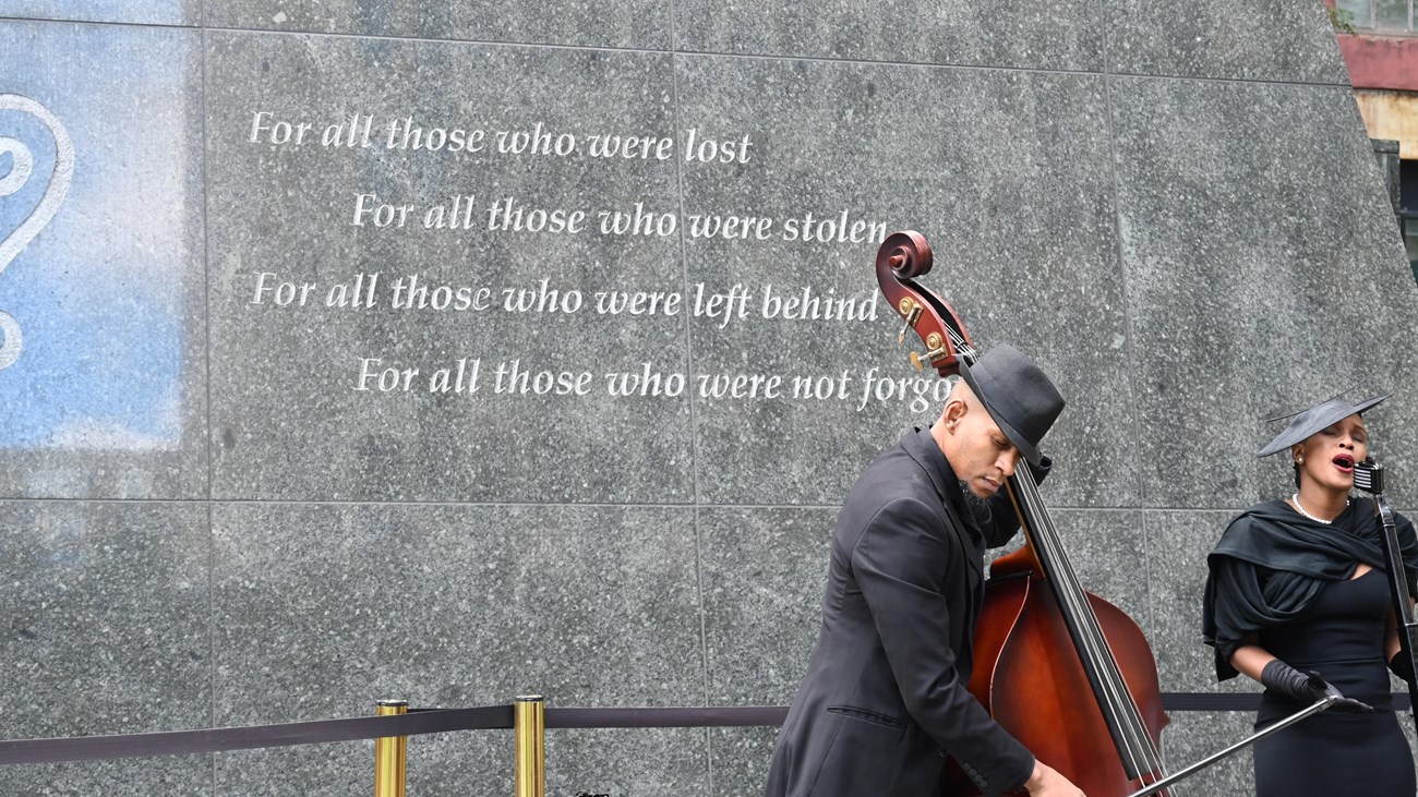 A man plays the bass and a woman sings in front of the monument. 