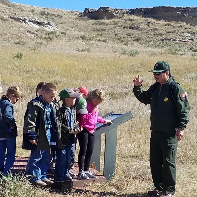 Male ranger gestures towards group of 10 young students on a grassy trail