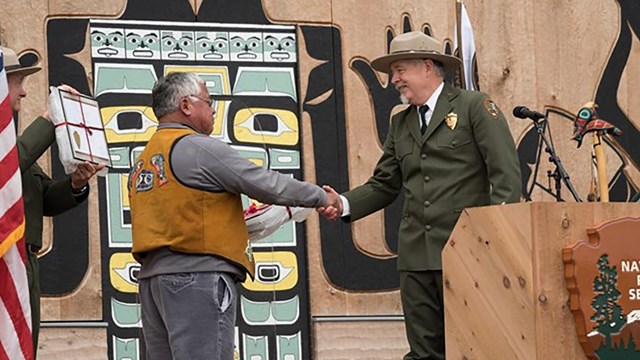 Two men shake hands, one is dressed in native traditional clothing, the other is in an NPS uniform