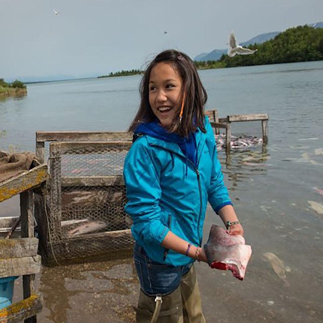 A young girl stands on the edge of water holding a clean, cut piece of fish.