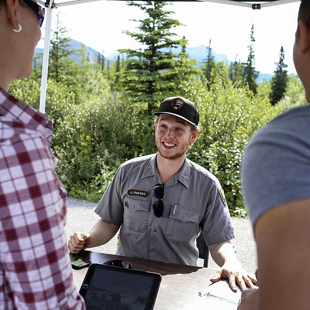 a ranger in uniform speaks to two visitors in a booth at an event in Denali national park