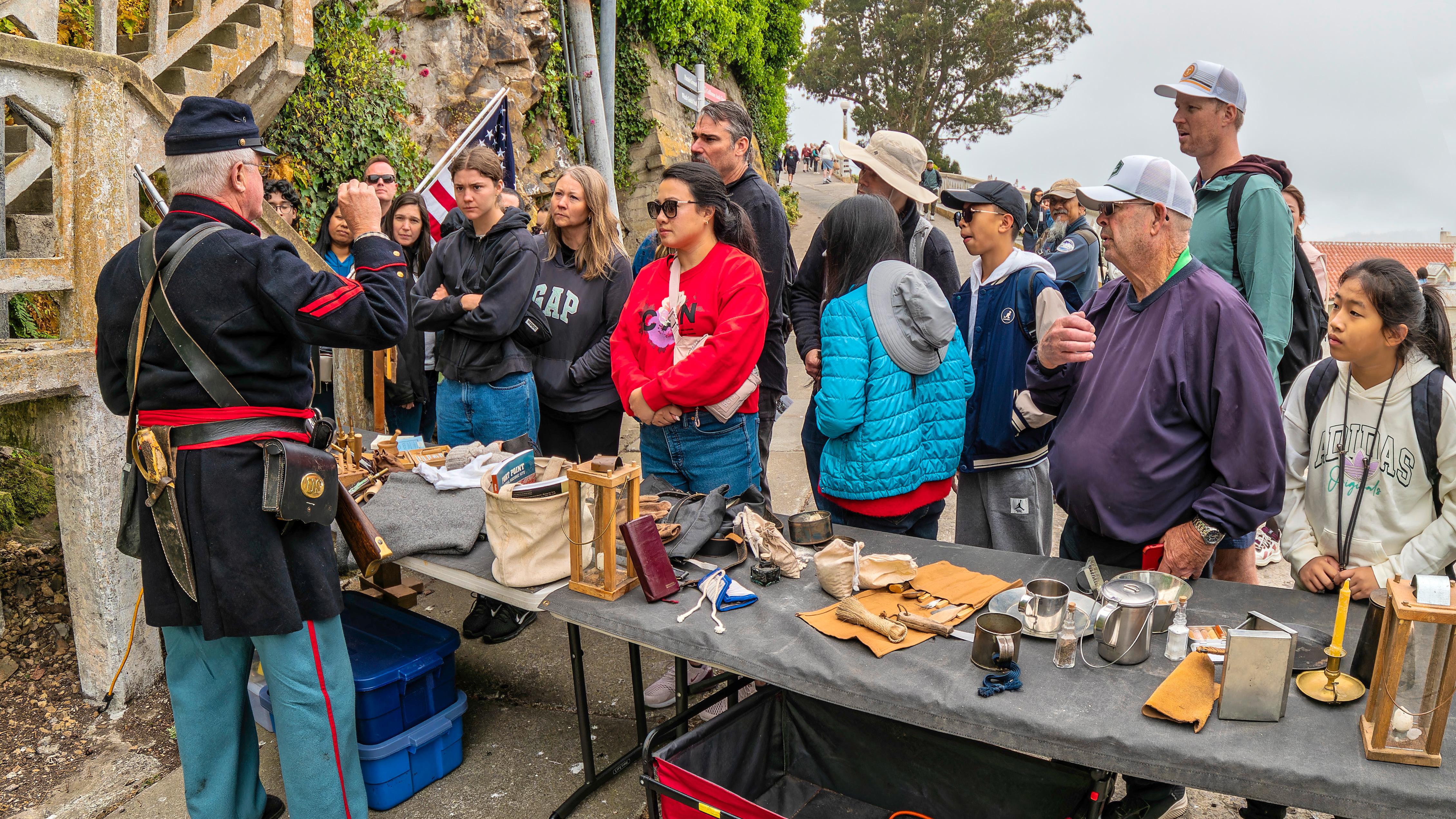 Man in blue Civil War uniform talks to a crowd. Table displays various historic objects.