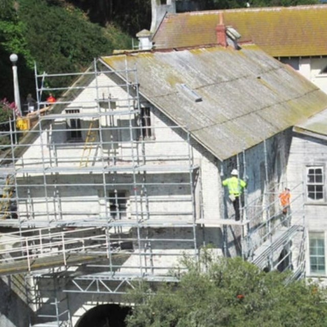 Construction workers in hardhats stand on scaffolding surrounding two-story historic building.