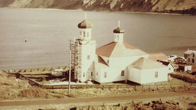 a church beside a coastal mountain is renovated with a scaffold structure on its steeple.