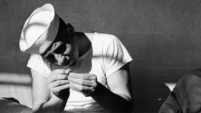 a historic scene of a sailor in a white cap squinting and threading a needle.