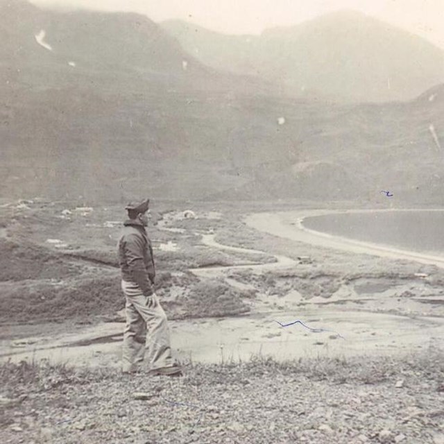 A man stands on a hill above a treeless river valley and distant buildings