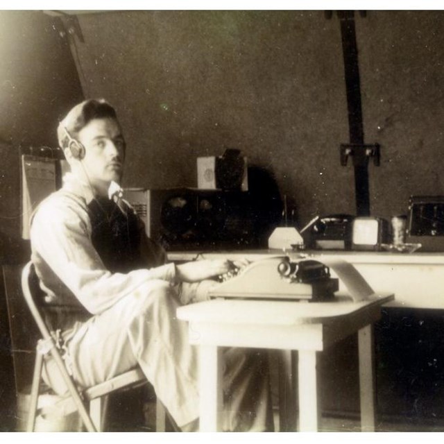 Black and white photo of man seated at typewriter, wearing radio headset, in an office. 