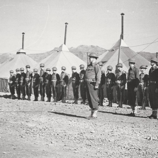 Sepia-toned photo of men in uniform in front of tents, on parade ground. 