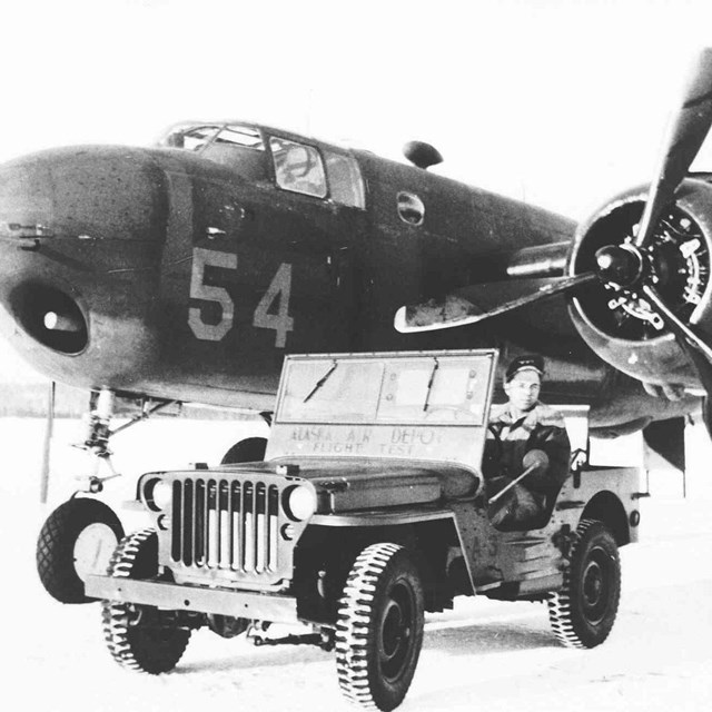 Black and white photo of B25 airplane and jeep with man in it. 