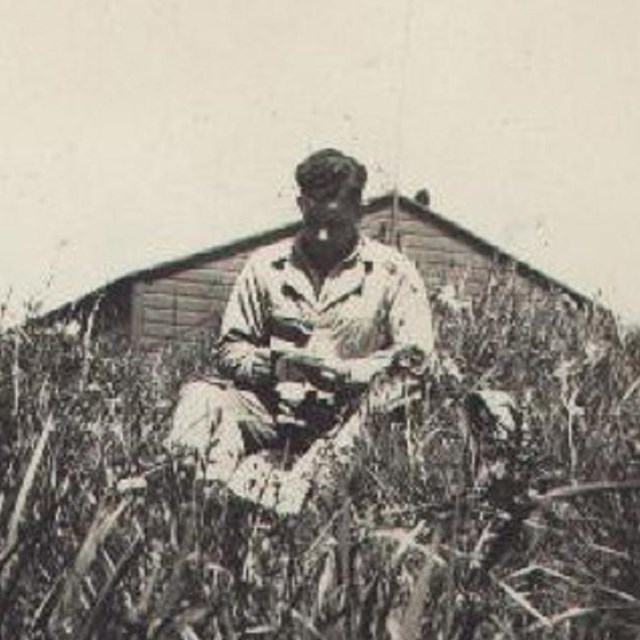 Man reading letter in meadow, with wooden building and sky in background. 