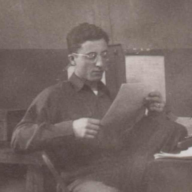 in a historic scene, a young man with glasses reads at a messy desk.