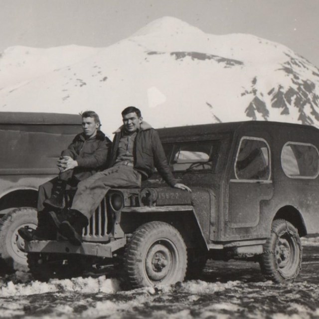 in a historic scene, two men sit on the hood of a jeep in front of a snow-covered mountain.