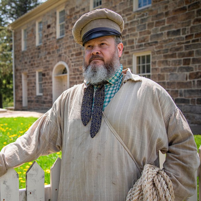 A ranger in period clothing stands in front of a stone tavern.