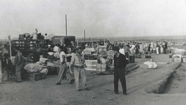 Two men carry a large crate. Behind them are crowds of people unloading suitcases off trucks