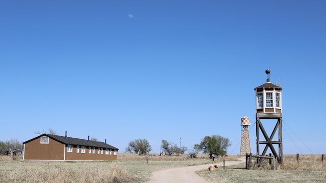 A road leads between a barrack and a guard tower with trees and a water tower on the horizon