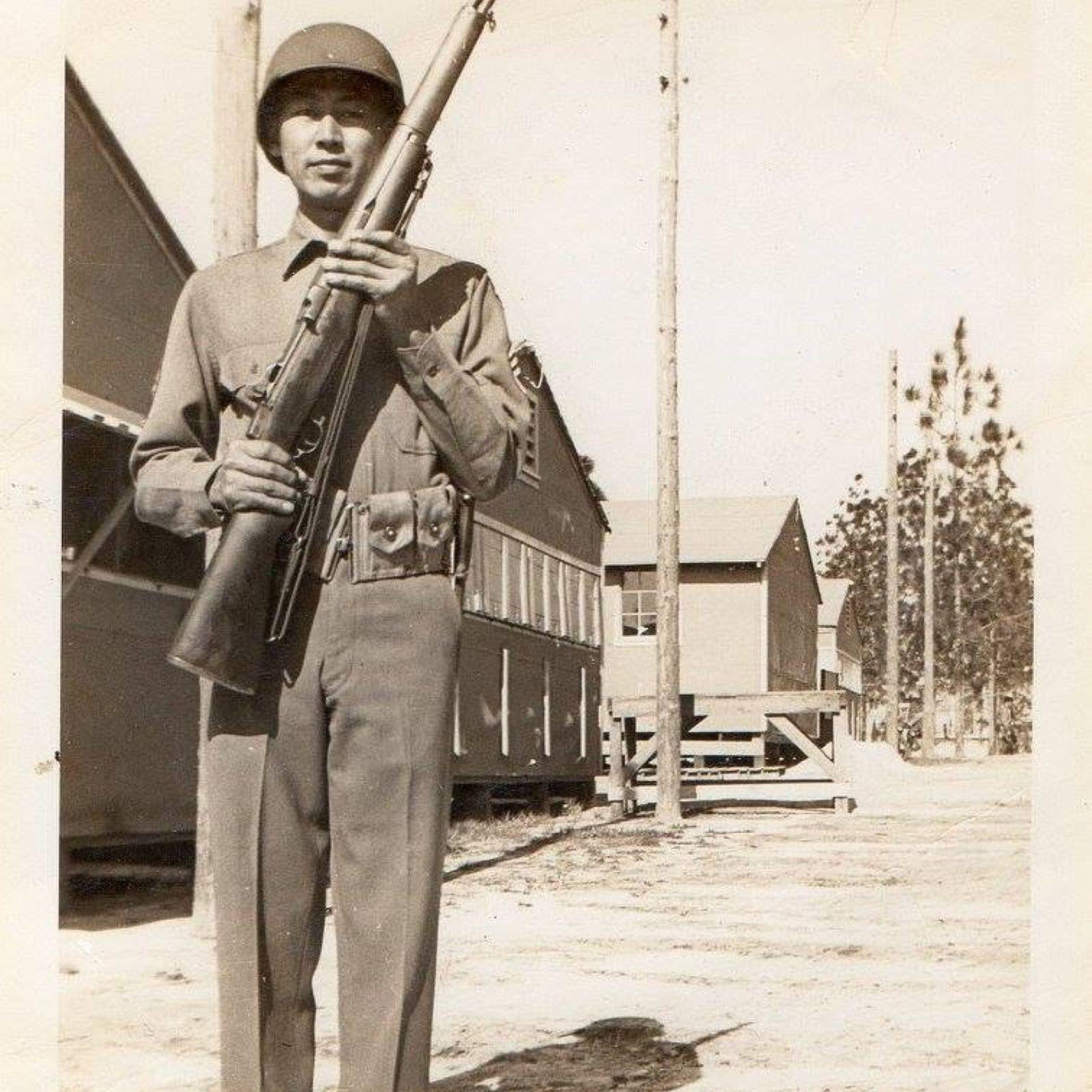 A man in an Army uniform stands with his firearm in front of a barrack.