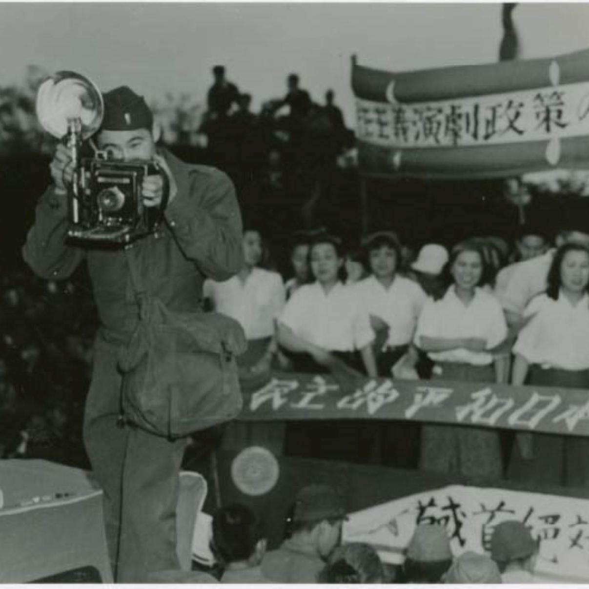 A man in an Army uniform takes a photo with a 1940s era camera. He is standing in front of a crowd.