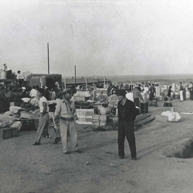 Two men carry a large crate. Behind them are crowds of people unloading suitcases off trucks
