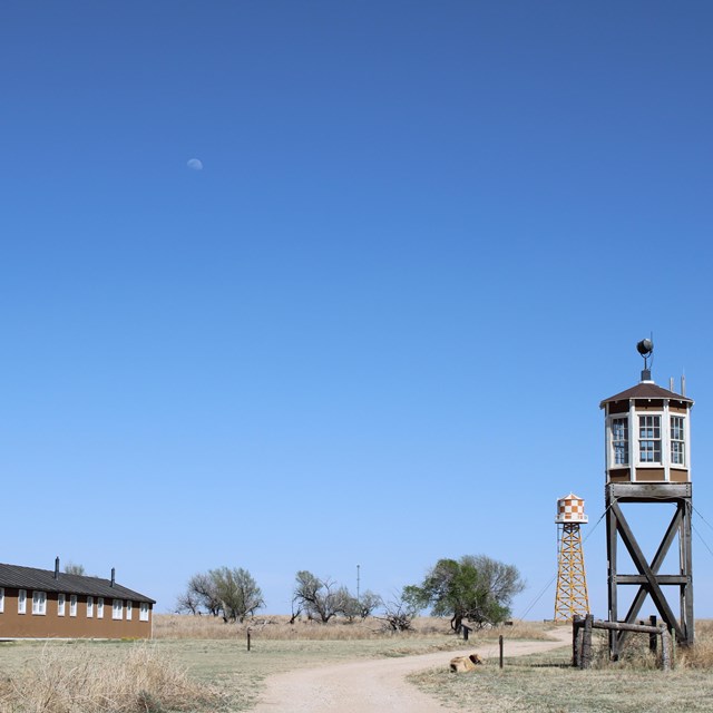 A road leads between a barrack and a guard tower with trees and a water tower on the horizon