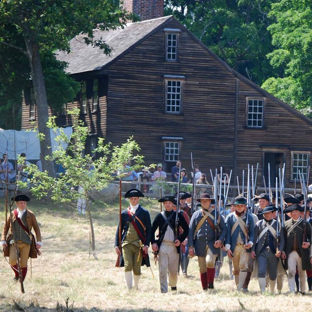 A line of militiman carrying muskets marches a field in front of a tw-story brown wood building 
