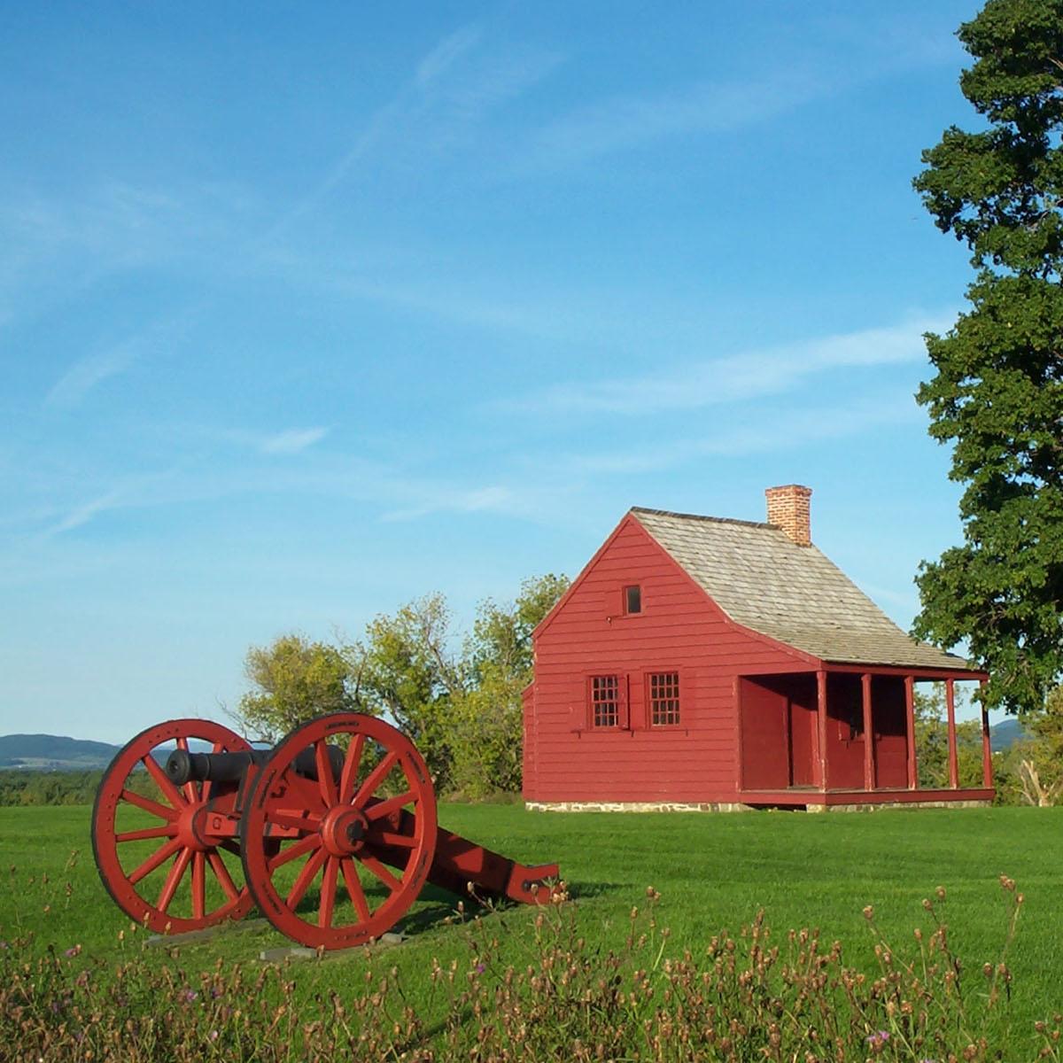 A cannon with red wheels stands on the green grass in front of the red Neilson house.