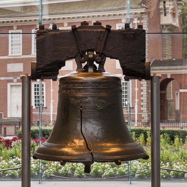 The Liberty Bell suspended between two posts. Independence Hall can be seen through the window.