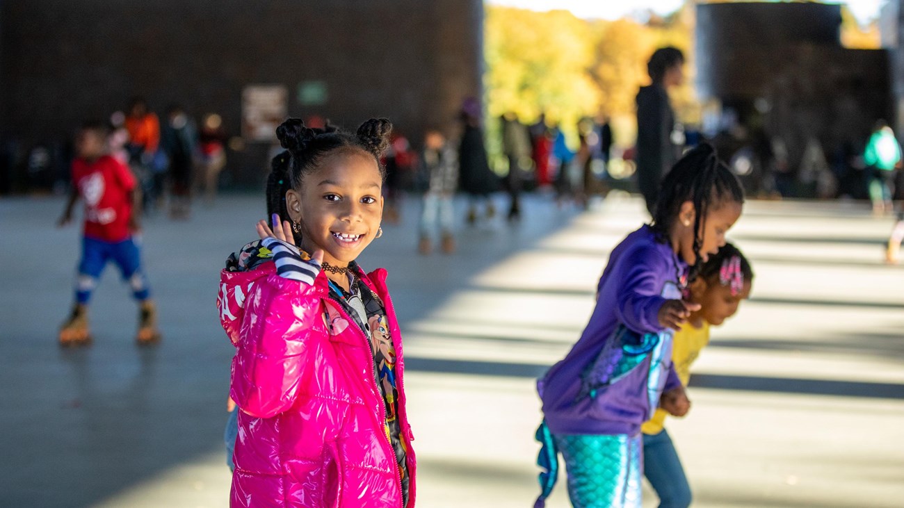 A young girl in a pink jacket waves and smiles at the camera as she skates by